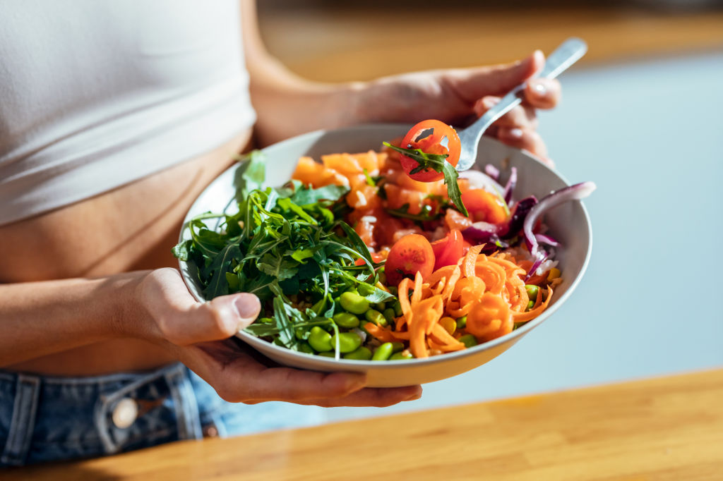 Manos sosteniendo un plato con ensalada de hojas verdes, tomate, zanahoria, edamame y salmón